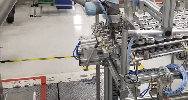 Línea de producción automatizada en una fábrica. Específicamente, se observa un sistema de embalaje que incluye un brazo robótico y una cinta transportadora con varias latas